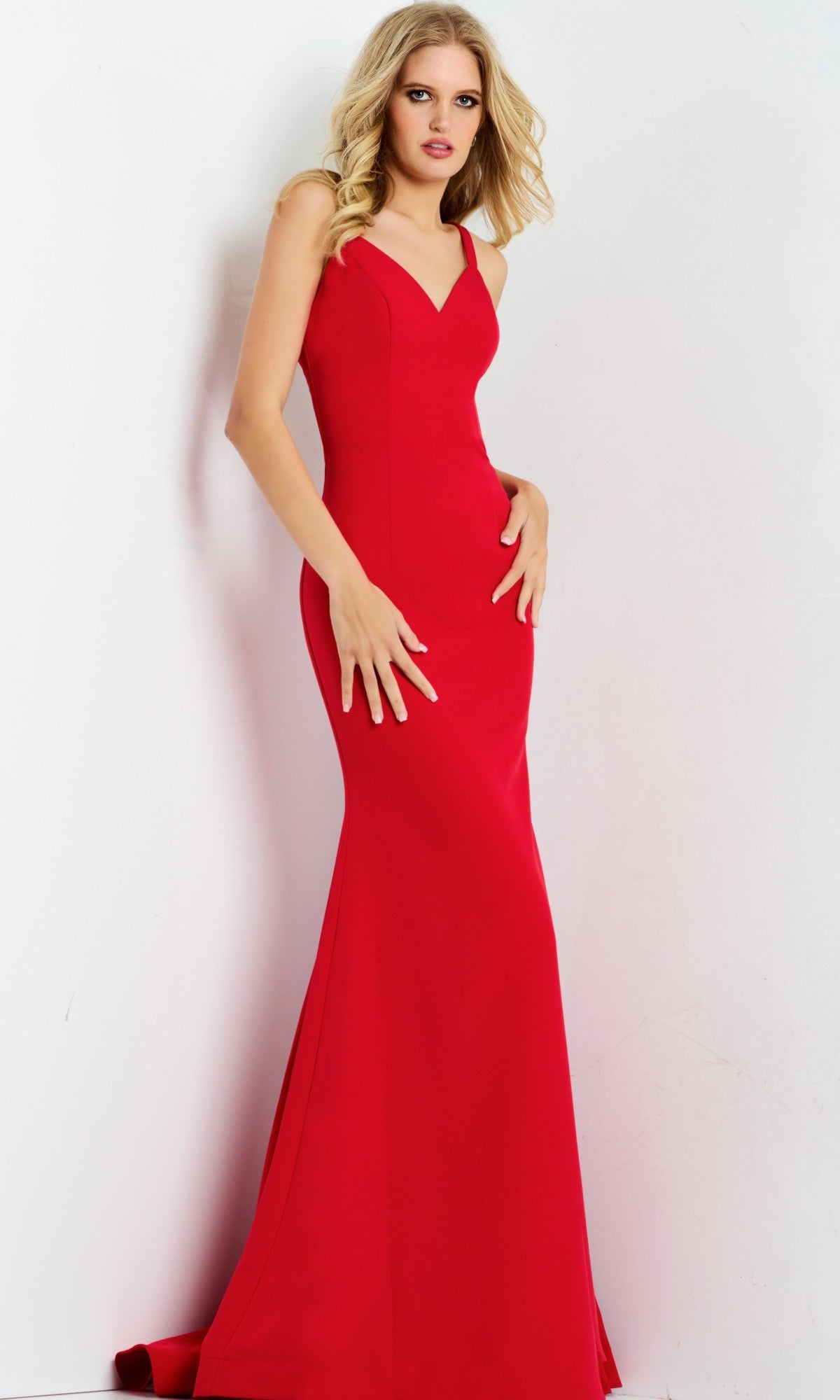 A woman stands against a white wall wearing the Jovani 49861 Long Prom Dress, a fitted, sleeveless red gown with a V-neckline, strappy open back, and flared hem.